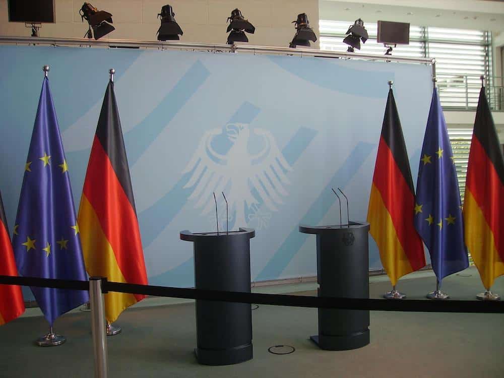 Deutschland und Europafahnen stehen vor einem Deutschland-Plakat mit dem deutschen Adler drauf. Davor stehen 2 Pulte, an denen Regierungssprecher normal ihre Reden halten.