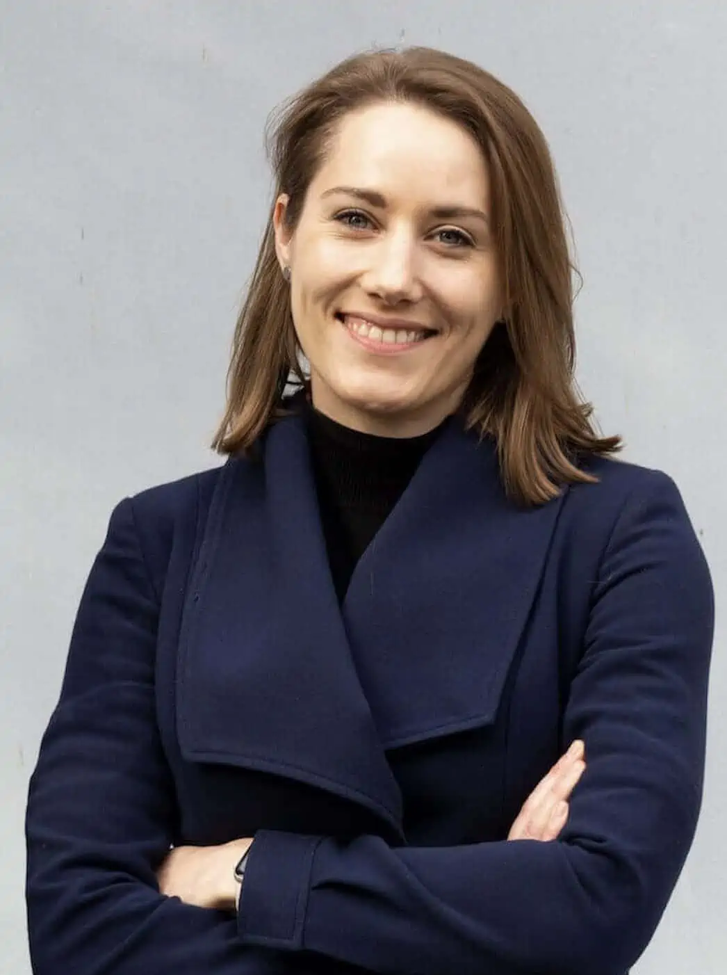 Nina Jameson steht mit verschränkten Armen vor einem grauen Hintergrund und lächelt in die Kamera
