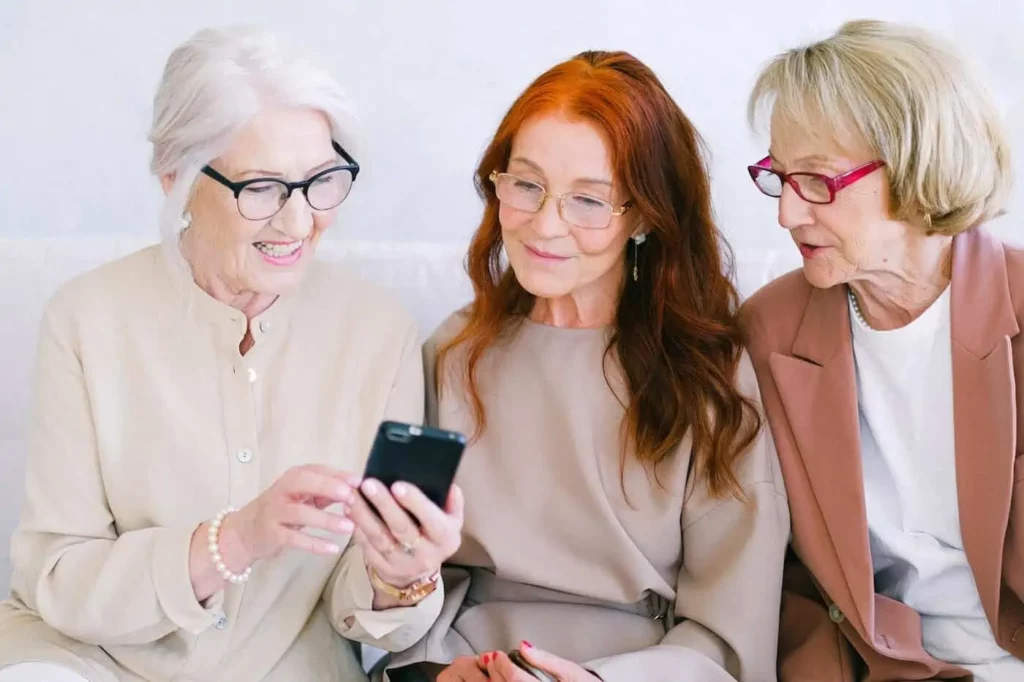 Drei ältere Frauen sitzen zusammen auf einem Sofa. Eine Dame hat ihr Handy in der Hand und zeigt den anderen zwei etwas darauf. wir sehen nur den hinteren Teil des Handys.