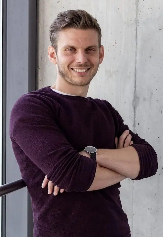 Tobias Roppelt steht mit verschränkten Armen vor einer Betonwand und lächelt in die Kamera. Er hat kurze, straßenköterblonde Haare, einen weinroten Pullover an und die Ärmel bis zur Mitte der Unterarme hochgekrempelt.