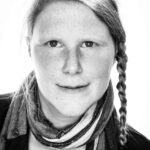 Ein schwarz-weißes Portrait von Daniela Schubert. Daniela lächelt in die Kamera. Daniela trägt einen Schal und einen seitlich geflochtenen Zopf.