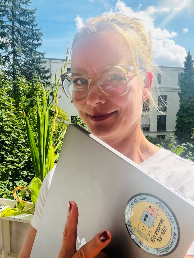 Nora Urru steht im Freien, mit ihrem Laptop in der Hand. Ihr Laptop hat den digitalen Barrierefreiheit-Sticker darauf!