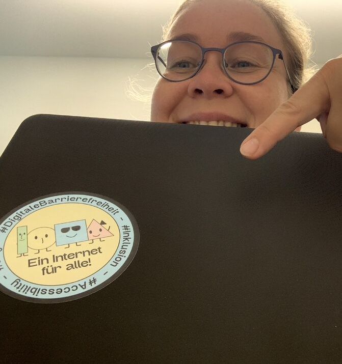 Katrin Zinke mit ihrem Laptop in der Hand. Ihr Laptop hat den digitalen Barrierefreiheit-Sticker darauf!